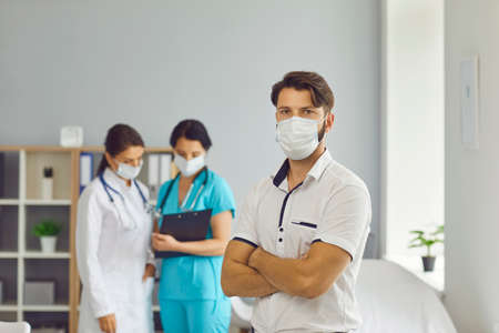 Portrait of a young male patient in a medical mask on the background of two female doctors. Man was...