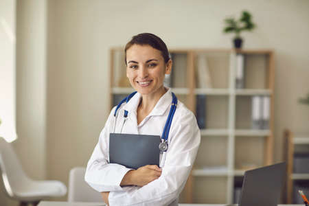 Young smiling woman doctor therapist in white traditional medical uniform standing and looking at camera over medical clinic office at background. Young professional doctor conceptの写真素材