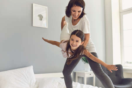 Happy mother and her preschool daughter have fun playing together on the bed at home. Woman lifts baby and she seems to be flying in moms arms in bedroom with modern interior.の写真素材