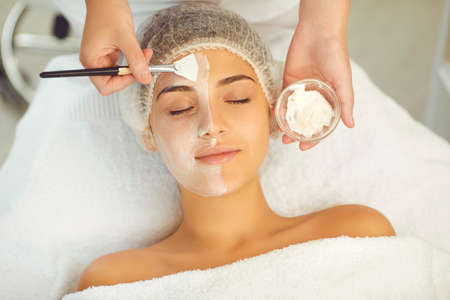 Facial treatment and skincare concept. Hands of dermatologist putting moisturizing mask to young relaxing womans face during skincare procedure in spa salon, top viewの写真素材