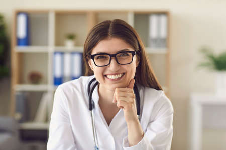 Professional medical service. Head shot of friendly female doctor looking at web camera during online video consultation with client. Profile picture of smiling general practitioner in hospital officeの写真素材