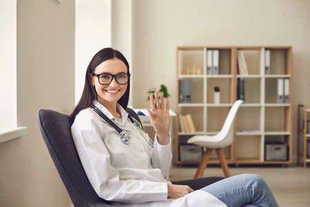 Young smiling woman doctor therapist in medical uniform sitting, looking at camera and greeting...