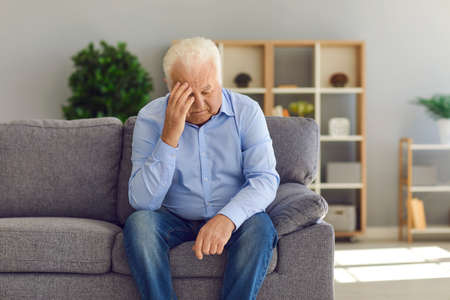 Tired, depressed senior man sitting on couch in living room feeling hurt and lonely. Aged,...