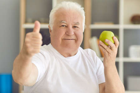 An apple a day keeps the doctor away. Eating healthy food and staying young in old age concept. Headshot of happy senior man in T-shirt holding fresh green apple and giving thumbs-up looking at cameraの写真素材