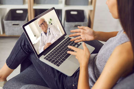 Young Asian woman using laptop computer at home and sharing her health problem with serious experienced senior doctor during online video consultation. Concept of telemedicine and e-healthの写真素材