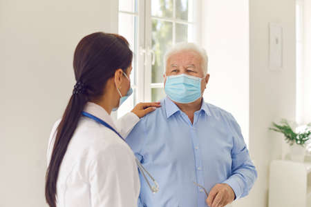 Senior man and young female doctor in medical face masks talking in hospital office. Physician in...