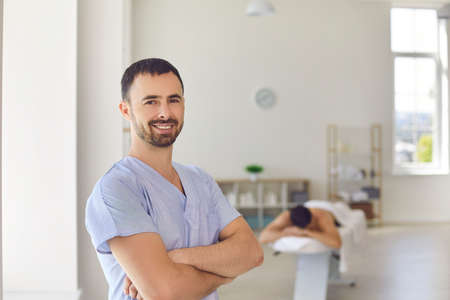 Portrait of smiling young professional masseur or physiotherapist looking at camera standing arms...