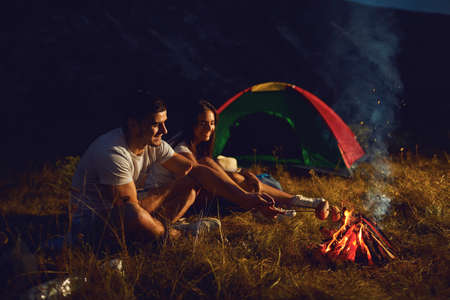 Young couple tourists on camping night cook bread by campfire. Happy people and bonfire in the...