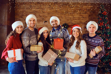 Happy friends in Santas hats give presents at Christmas. A group of young people in a room with a...