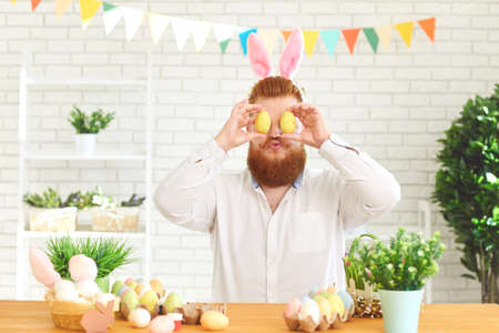 Happy easter. A funny bearded fat man decorates eggs while sitting at a table with Easter decor in the background.の写真素材
