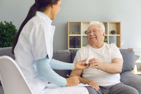 Female doctor visiting her cheerful senior male patient at home, prescribing treatment, giving him...