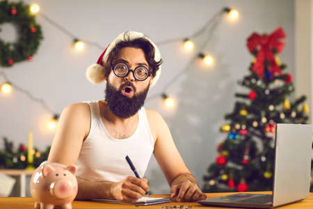 Funny young man in hat and glasses looking at camera, sitting at desk with laptop and piggy bank, writing letter to Santa or making list of wishes, dreams, plans and goals for upcoming year in notepadの写真素材