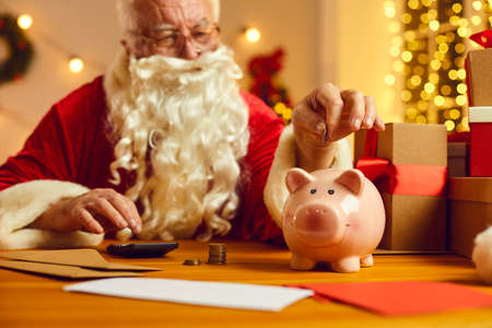 Santa Claus putting coins in piggy bank while sitting at desk with presents, calculator, letters and...