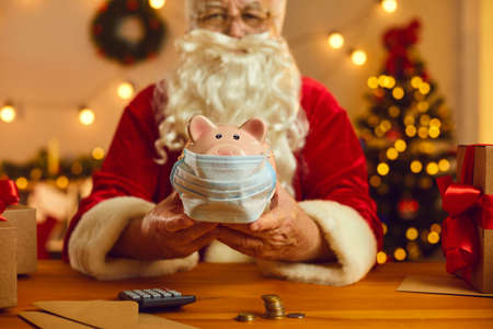 Santa Claus at desk with money and calculator holding piggy bank in medical face mask. Healthcare financing and medical care sponsorship, helping fight Covid 19, donation, fundraising, charity conceptの写真素材
