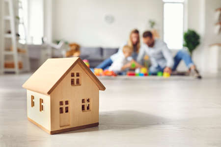 Close-up of miniature detached house on floor at home with happy family playing with little child in background. Mortgage, real estate business, buying property and confidence about future conceptsの写真素材