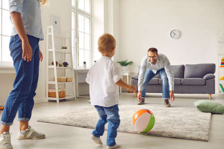 Happy family staying at home, having fun together and engaging their small child in sport games. Young dad with little son playing football with inflatable ball in living-roomの写真素材