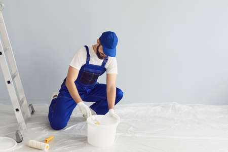 Bearded male painter in uniform taking liquid pigment from bucket against gray wall during renovation works in modern apartmentの写真素材