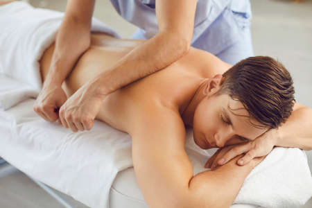 Young man enjoying professional body treatment in spa salon. Relaxed client lying on bed with eyes closed and getting remedial body massage done by male massagist in health center or massage roomの写真素材