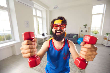 Happy crazy beginner athlete with weak arm muscles who decided to start fitness training and exercise with dumbbells is looking at camera and laughing, motivating you to do sports and keep fit tooの写真素材