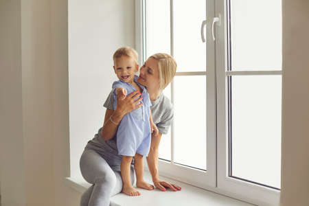Happy young mommy hugging cute toddler boy sitting by large window with newly installed window pane...