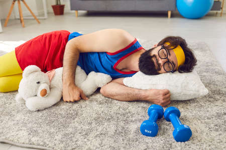 Happy, funny, relaxed, lazy man in activewear who never started exercising or working out, sleeping peacefully on floor at home, hugging a teddy bear, with dumbbells beside. Lack of motivation conceptの写真素材