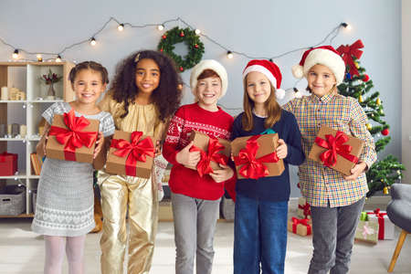 Group of happy smiling diverse children holding Christmas presents tied with beautiful red bows and looking at camera standing in cozy festively decorated living-room during fun party at homeの写真素材