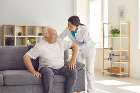 Female nurse assisting happy senior patient with daily activities in nursing home. Young woman helping aged man to stand up from couch in room of modern hospital or geriatric rehabilitation centerの写真素材