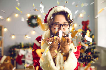 Christmas and New Year. Cheerful young man in Santa suit and hat blows confetti with gold glitter...