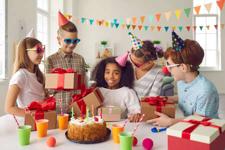 Group of children in funny party hats and sunglasses giving birthday presents to happy smiling African-American girl sitting at festive table with cake and juice during fun celebration at home.の写真素材
