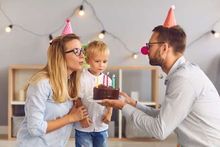 Happy, loving young parents in funny party hats giving chocolate cake with 5 burning candles to their shy little birthday boy during quiet celebration at homeの写真素材
