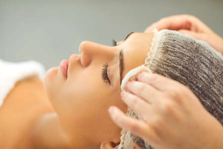 Beauticians hands are wiping the womans face with a cotton pad in a beauty salon. Young female client lies in a medical cap with her eyes closed and prepares for cosmetic procedures.の写真素材