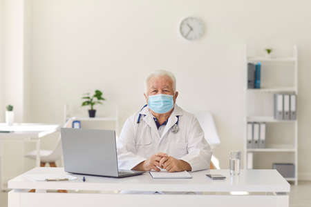 Portrait of a serious senior doctor in protective mask and medical gown who sits with his desk at...