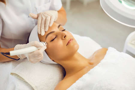Relaxed young woman with eyes closed getting procedure of vacuum facial cleaning from cosmetologist in gloves in beauty spa salon. Facial treatment, skincare, cosmetology conceptの写真素材