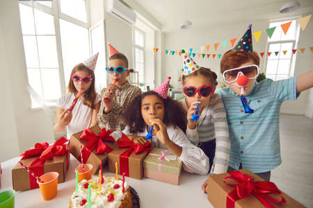 Group of happy diverse children in party hats and funny sunglasses blowing noisemakers and looking at camera while having fun during birthday celebration at homeの写真素材