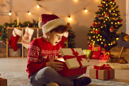 Boy in Santa holiday hat and red festive sweater feeling surprised opening holiday Christmas gift box with miracle inside over Christmas decorated room at background. New Year magic conceptの写真素材