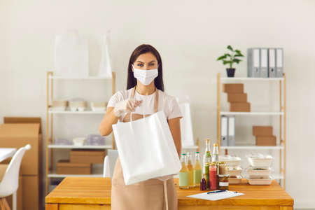Responsible takeaway cafe worker in face covering mask and gloves holding paper bag with fresh lunch ready to be delivered to client. Safe meal delivery service and taking care of customers conceptの販売画像