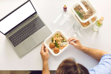 Top view of cropped busy worker sitting at office desk with laptop computer with blank white mockup copyspace screen, enjoying lunch break and eating healthy meal ordered on food delivery websiteの写真素材