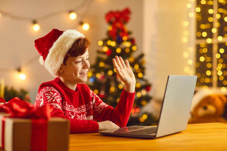 Online Xmas party. Happy smiling child sitting in cozy decorated room at home, waving hand at screen...