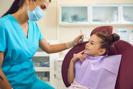 Happy, curious little patient trusting her doctor. Cute 6 - 8 year old girl sitting in dental clinic chair and talking to friendly female dentist in medical mask ready to examine teeth of young clientの写真素材