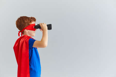 Little boy in a mask cloak of a superhero looks through binoculars towards a free space for text standing in profile on a gray background.の写真素材
