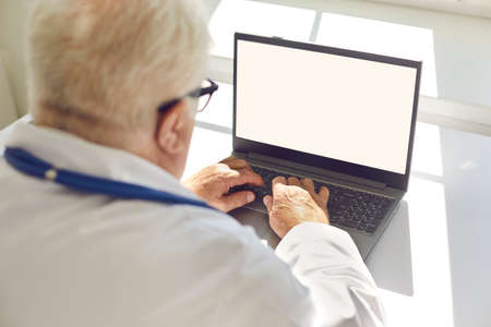 Senior man sitting in a bright room behind a laptop and typing on the keyboard. Man uses a laptop to communicate or read the morning news. Place for text. Close up.の写真素材
