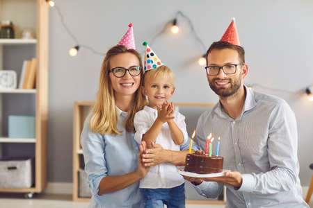 Portrait of happy, loving, young parents and their little birthday boy, wearing funny party caps, holding chocolate cake, looking at camera and smiling during quiet party at homeの写真素材