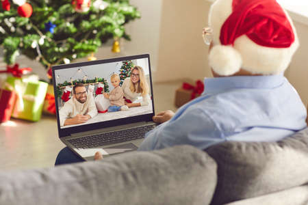 Senior man grandfather in Santa festive hat sitting on sofa and communicating with family online...