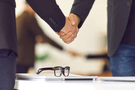 Close up of two businessmen shaking hands over a negotiating table in the office. There is a notebook and glasses on the table. Partners corporate relationship concept.の写真素材