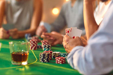Hands of people friends with drinks and playing cards betting and playing poker on table at home together, selective focus. Gambling, casino at home, board games, playing cards conceptの写真素材