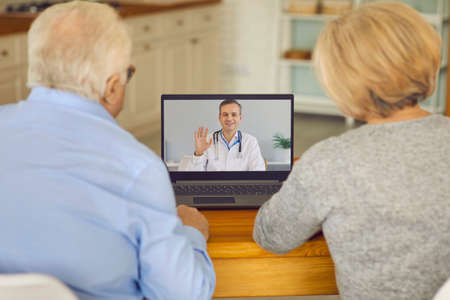 Medical online consultation. Back view of a senior couple sitting at a table in front of a laptop at...