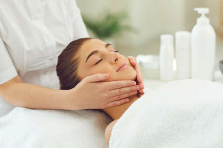 Young woman getting procedure of relaxing facial massage from hands of masseur in beauty spa salon. Rejuvenating and lifting massage in cosmetology conceptの写真素材