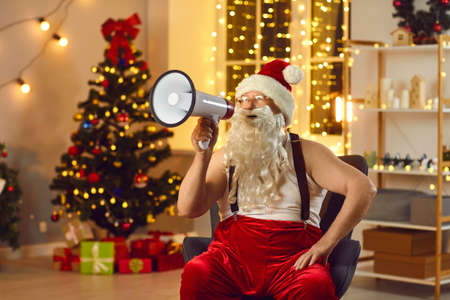Funny Santa Claus in suspender pants sitting in armchair with megaphone in hand announcing the start of big New Year and Christmas sales and discounts, promising amazing deals, gifts and presentsの写真素材