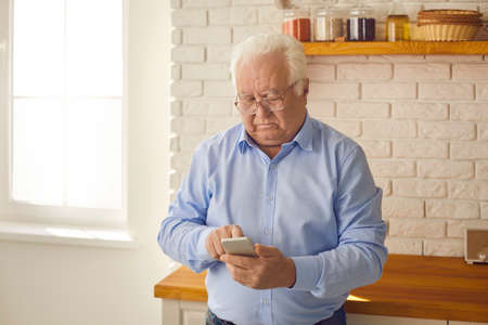 Serious senior man in glasses standing in the kitchen and using online shopping app, texting or...