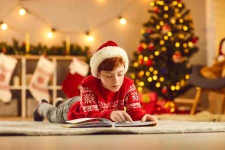 Concentrated boy in red Christmas clothing and hat lying on floor and reading book alone over...